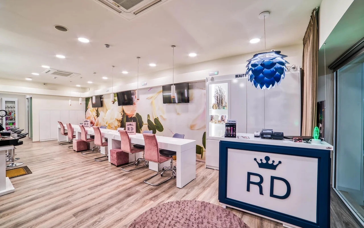 Ein hell-weisser Salon mit einer langen Reihe von Maniküre-Tischen und altrosa Stühlen, einem sanften floralen Wandbild, hellem Holzboden und einer Rezeptionstheke mit einem 'RD'-Kronenlogo.