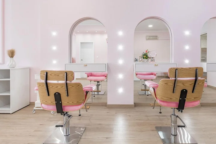 Heller Coiffeursalon in Zart-Rosa mit Frisierstühlen aus Holz und rosa Stoffbezug vor zwei bogenförmigen, punktbeleuchteten Spiegelnischen, weissen Schränken und Boden in heller Holzoptik.