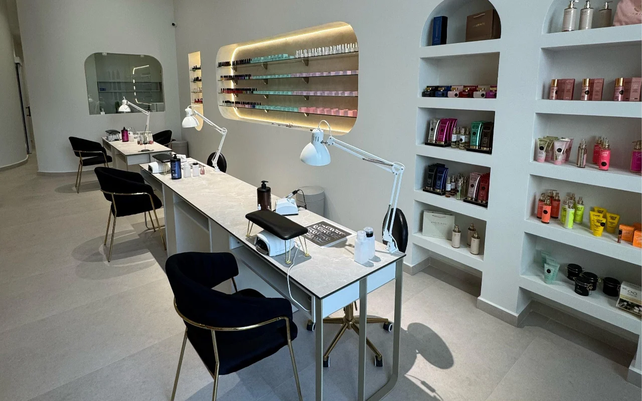 Ein weisses Nagelstudio mit Maniküretischen mit Marmorplatten und weissen Gelenklampen, schwarzen Samtstühlen auf goldenen Beinen, und gewölbten Wandnischen mit Skincare-Produkten und einem hintergrundbeleuchteten Nagellack-Regal.