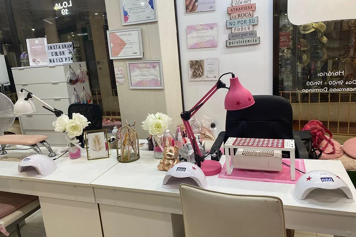 Maniküre-Station eines Nagelsalons mit weissen Tischen, einer kräftig rosa Lampe und LED-Lampen, weissen Rosen und gerahmten Zertifikaten an der Wand, mit dem Namen Color Nails PT auf der Scheibe neben den Öffnungszeiten sichtbar.