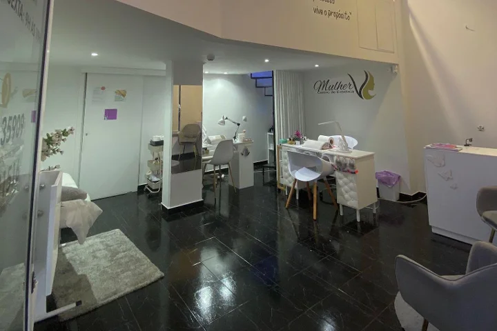 Innenraum eines Kosmetikstudios: dunkler polierter Fliesenboden, weisser Maniküre-Tisch und dezent beleuchtete Behandlungsbereiche, das Mulher-Logo an der Rückwand und eine Gesichtsbehandlungsliege hinter einer Glastrennwand sichtbar.