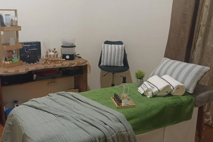 Gemütlicher Massageraum mit einer mit einem grünen Handtuch bezogenen Liege und aufgerollten weissen Handtüchern, einer Schale ätherischer Öle, Holz-Massagewerkzeugen auf einem rustikalen Beistelltisch und einem Hocker neben einer durch einen Vorhang abgetrennten Ecke.