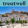Sonniger Blick über Ibiza-Stadt, mit weiss getünchten Gebäuden, die zur Kathedrale und den alten Festungsmauern von Dalt Vila auf dem Hügel hinaufsteigen. Eine von Bäumen gesäumte Hafenpromenade zieht sich unter einem klaren blauen Himmel durch den Vordergrund.