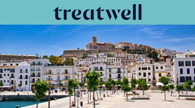 Sonniger Blick über Ibiza-Stadt, mit weiss getünchten Gebäuden, die zur Kathedrale und den alten Festungsmauern von Dalt Vila auf dem Hügel hinaufsteigen. Eine von Bäumen gesäumte Hafenpromenade zieht sich unter einem klaren blauen Himmel durch den Vordergrund.