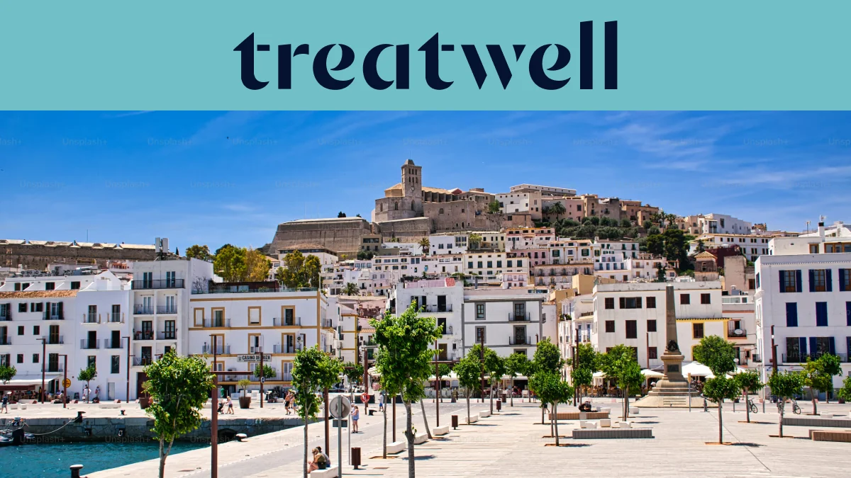 Sonniger Blick über Ibiza-Stadt, mit weiss getünchten Gebäuden, die zur Kathedrale und den alten Festungsmauern von Dalt Vila auf dem Hügel hinaufsteigen. Eine von Bäumen gesäumte Hafenpromenade zieht sich unter einem klaren blauen Himmel durch den Vordergrund.