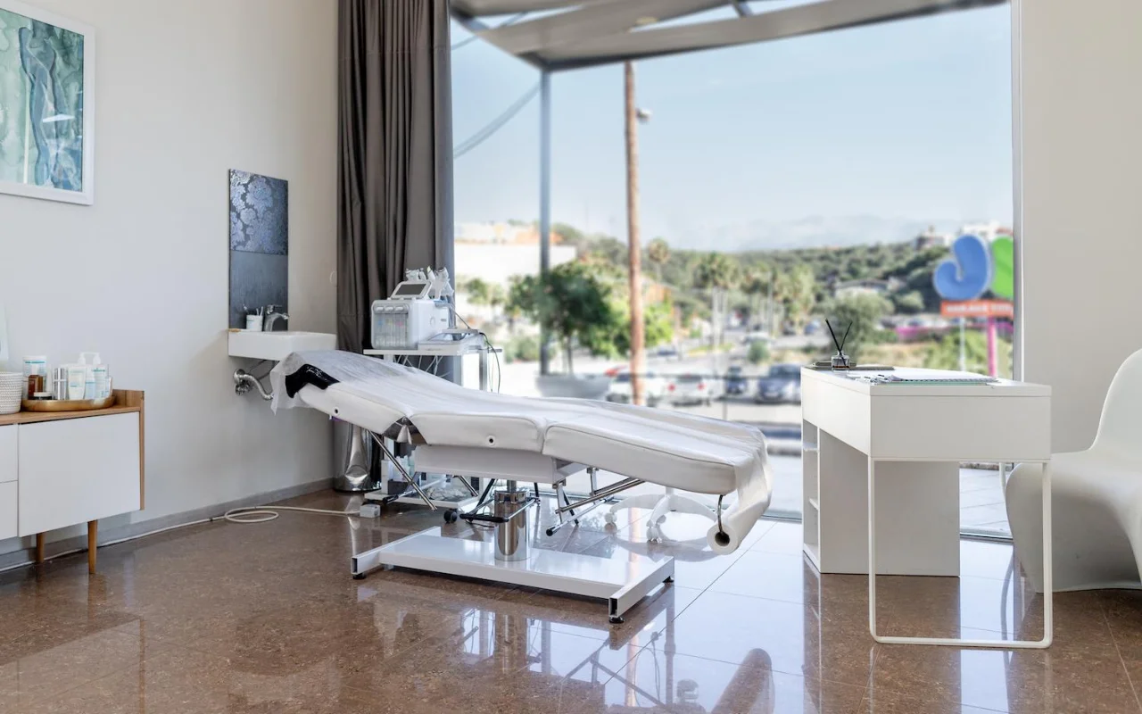 Ein sauberer weisser Behandlungsraum mit einer verstellbaren Beauty-Liege, Skincare-Geräten auf einem Wagen, gerahmten Kunstwerken und einem grossen Fenster mit Blick auf Bäume, eine Strasse und ferne Hügel.