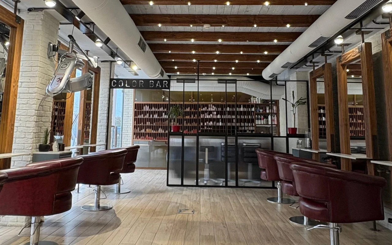 Ein Coiffeursalon mit freiliegenden Holzdachbalken und weiss getünchtem Ziegelstein, weinroten Lederstyling-Stühlen und einem verglasten Farbraum mit Regalen voller Produkte hinter einem 'Color Bar'-Schild.