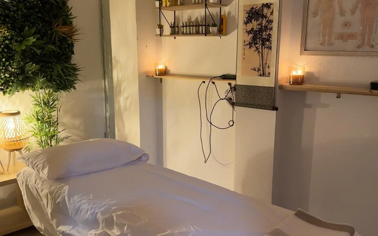 Ein schwach beleuchteter Behandlungsraum mit einer in weissem Leinen bezogenen Massageliege. Angezündete Kerzen stehen auf hölzernen Wandregalen neben einer Bambus-Tuschzeichnung und einer gerahmten Akupunktur-Tafel, mit einer Grünpflanze und einer leuchtenden Korblampe in der Ecke.