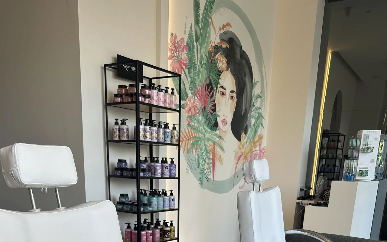 Eine Studio-Ecke mit zwei weissen verstellbaren Behandlungsstühlen, einem schwarzen Produkt-Regal, einem hintergrundbeleuchteten Spiegel und einem floralen Wandbild eines Frauengesichts, umrahmt von tropischem Laub.