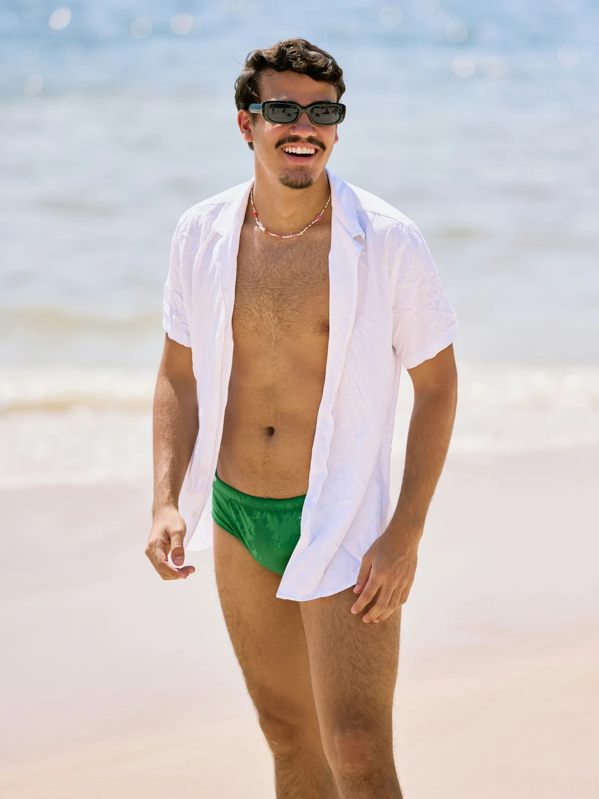 Ein lächelnder Mann steht an einem sonnigen Tag am Strand, er trägt eine grüne Badehose, ein offenes weisses Hemd und eine schwarze Sonnenbrille