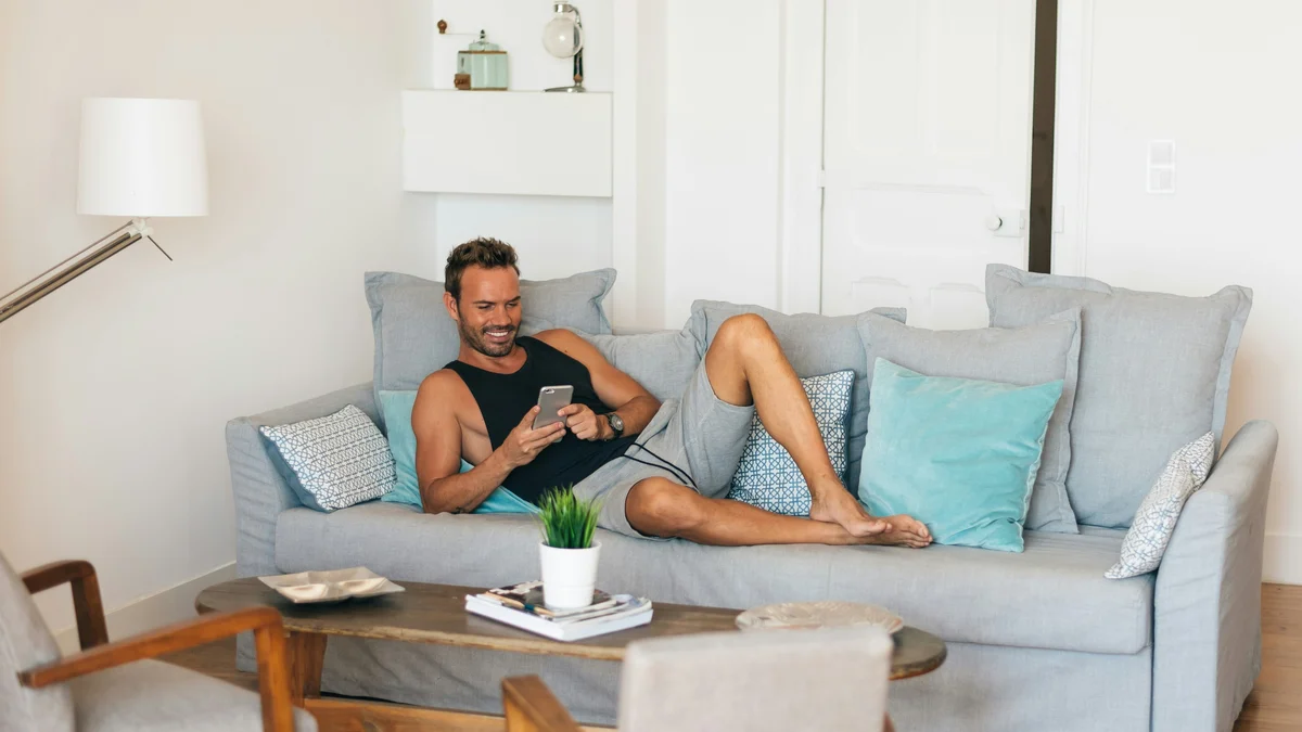 Ein Mann entspannt mit hochgelegten Füssen auf seinem Sofa und lächelt, während er auf sein Handy schaut; er trägt ein schwarzes Trägershirt und eine lockere, weiche graue Shorts