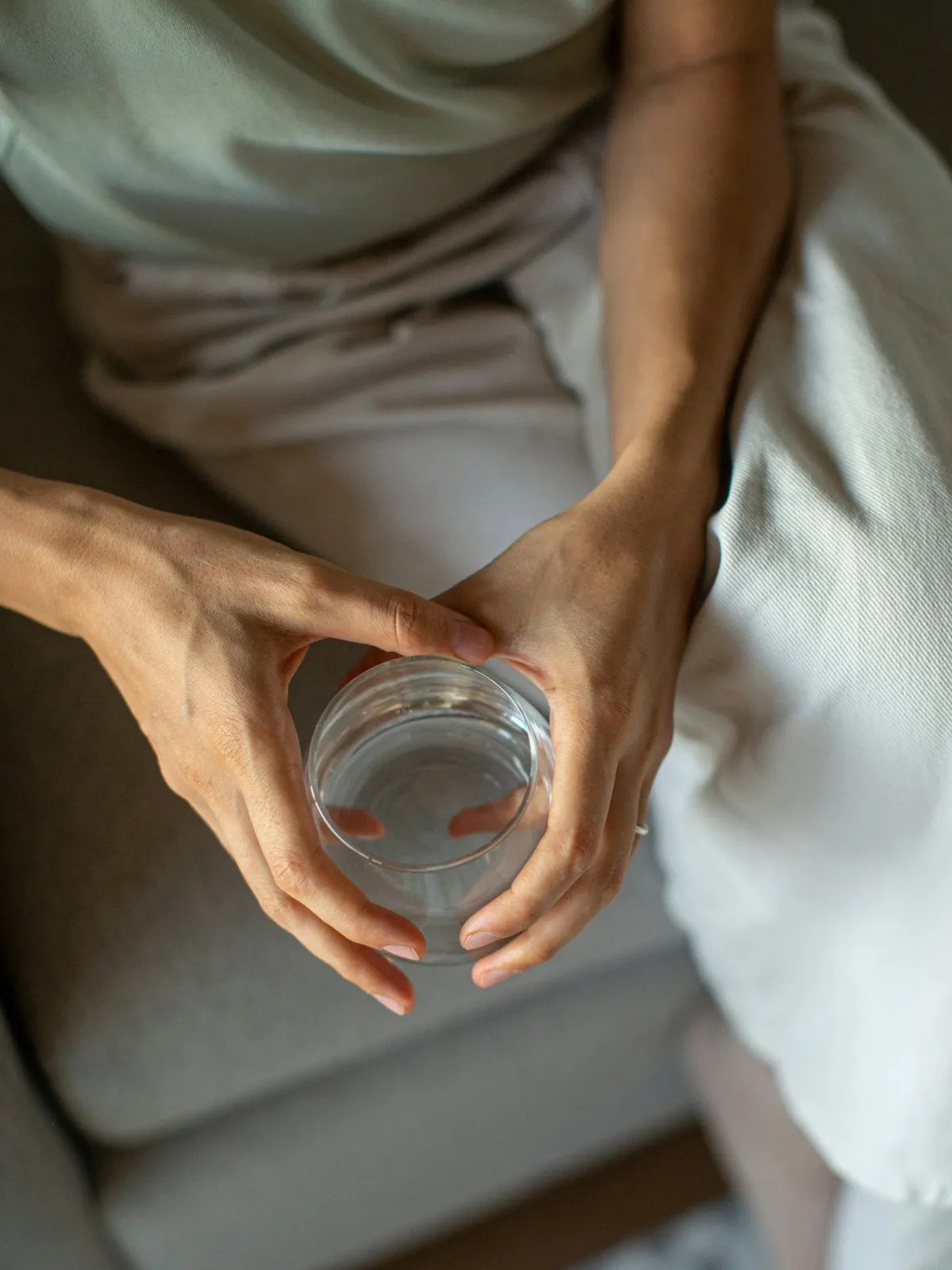 Aufsicht auf eine Person in lockerer Kleidung, die auf einem grauen Sofa sitzt und ein Glas Wasser mit beiden Händen hält